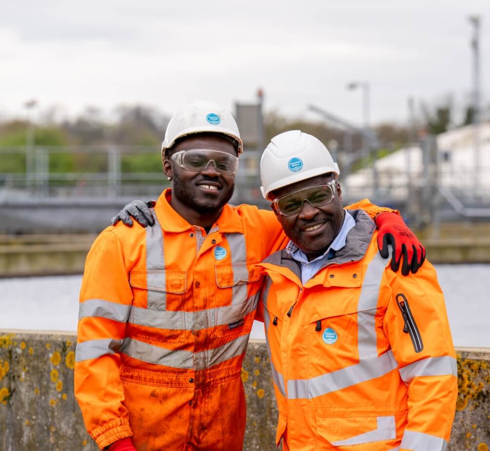 Two people with hard hats on and with arms around each other smiling at camera